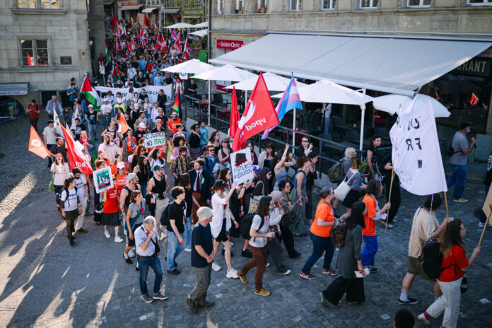 Vue en plongée d'une manifestation de rue dans ce qui semble être une ville suisse ou française. Des centaines de personnes marchent sur des pavés, brandissant des drapeaux rouges, des drapeaux palestiniens et un drapeau aux couleurs transgenres (rose, blanc et bleu). Des pancartes sont visibles, dont l'une indique "No Justice No Peace" et une autre "Droit à l'éducation". Des terrasses de restaurants bordent la rue.
