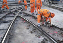 Initiative du chaos : qui peut rester, qui doit partir ? Plusieurs ouvriers du bâtiment vêtus de combinaisons orange haute visibilité et de casques de protection travaillent sur un chantier de réfection de rails de tramway. Au premier plan, un ouvrier se penche en avant et utilise un marteau-piqueur pour démolir le revêtement en béton autour des rails métalliques croisés. D'autres travailleurs sont visibles en arrière-plan sur le même chantier.