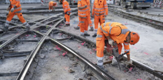 Plusieurs ouvriers du bâtiment vêtus de combinaisons orange haute visibilité et de casques de protection travaillent sur un chantier de réfection de rails de tramway. Au premier plan, un ouvrier se penche en avant et utilise un marteau-piqueur pour démolir le revêtement en béton autour des rails métalliques croisés. D'autres travailleurs sont visibles en arrière-plan sur le même chantier.