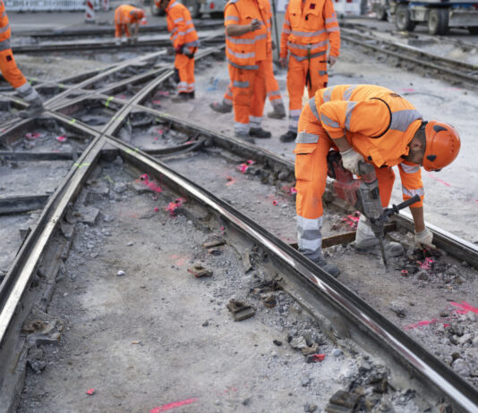 Initiative du chaos : qui peut rester, qui doit partir ? Plusieurs ouvriers du bâtiment vêtus de combinaisons orange haute visibilité et de casques de protection travaillent sur un chantier de réfection de rails de tramway. Au premier plan, un ouvrier se penche en avant et utilise un marteau-piqueur pour démolir le revêtement en béton autour des rails métalliques croisés. D'autres travailleurs sont visibles en arrière-plan sur le même chantier.