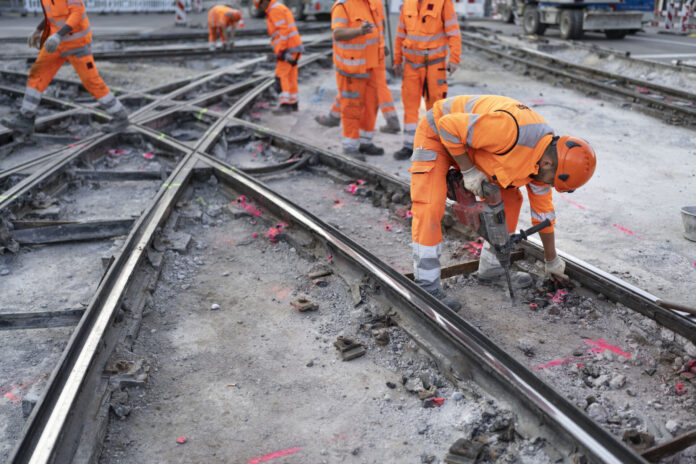 Plusieurs ouvriers du bâtiment vêtus de combinaisons orange haute visibilité et de casques de protection travaillent sur un chantier de réfection de rails de tramway. Au premier plan, un ouvrier se penche en avant et utilise un marteau-piqueur pour démolir le revêtement en béton autour des rails métalliques croisés. D'autres travailleurs sont visibles en arrière-plan sur le même chantier.
