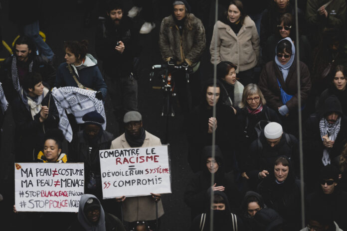 L'image montre un groupe de personnes participant à une manifestation à Lausanne après la mort de Marvin. Plusieurs individus tiennent des pancartes avec des messages contre le racisme et la violence policière. Les pancartes portent des inscriptions telles que "Combattre le racisme - Dévoiler un système sans compromis" et "Ma peau n'est pas un costume, ma peau n'est pas une menace #StopBlackFace #StopPoliceViolence". Les manifestants sont vêtus chaudement, suggérant que l'événement a lieu en hiver. L'atmosphère semble sérieuse et déterminée, avec des participants de divers âges et origines ethniques.