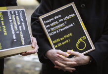 LAFE : Fribourg a un problème de recettes, pas de dépenses Deux personnes vêtues de manteaux sombres tiennent des pancartes artisanales à fond noir avec du texte en blanc et jaune. On peut lire sur la pancarte principale : « Couper dans les soins, les crèches, la formation ? NON à l'austérité ! NON au PAFE le 26 avril ! », accompagnée d'un pictogramme de bombe en jaune. La seconde pancarte, partiellement visible, affiche un message similaire.