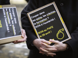 LAFE : Fribourg a un problème de recettes, pas de dépenses Deux personnes vêtues de manteaux sombres tiennent des pancartes artisanales à fond noir avec du texte en blanc et jaune. On peut lire sur la pancarte principale : « Couper dans les soins, les crèches, la formation ? NON à l'austérité ! NON au PAFE le 26 avril ! », accompagnée d'un pictogramme de bombe en jaune. La seconde pancarte, partiellement visible, affiche un message similaire.