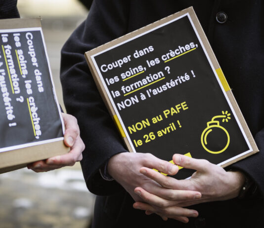 LAFE : Fribourg a un problème de recettes, pas de dépenses Deux personnes vêtues de manteaux sombres tiennent des pancartes artisanales à fond noir avec du texte en blanc et jaune. On peut lire sur la pancarte principale : « Couper dans les soins, les crèches, la formation ? NON à l'austérité ! NON au PAFE le 26 avril ! », accompagnée d'un pictogramme de bombe en jaune. La seconde pancarte, partiellement visible, affiche un message similaire.