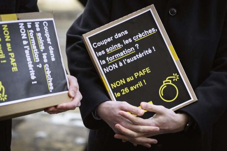 Deux personnes vêtues de manteaux sombres tiennent des pancartes artisanales à fond noir avec du texte en blanc et jaune. On peut lire sur la pancarte principale : « Couper dans les soins, les crèches, la formation ? NON à l'austérité ! NON au PAFE le 26 avril ! », accompagnée d'un pictogramme de bombe en jaune. La seconde pancarte, partiellement visible, affiche un message similaire.