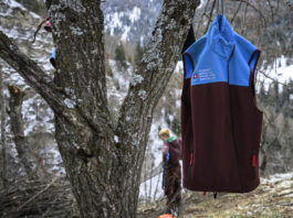 Affaiblissement du service civil : une attaque frontale de la droite Un gilet du Service civil suisse (bicolore bleu et bordeaux) accroché à un arbre en forêt, avec en arrière-plan une personne travaillant dans un paysage hivernal de montagne.