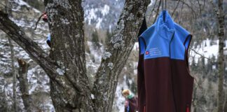Affaiblissement du service civil : une attaque frontale de la droite Un gilet du Service civil suisse (bicolore bleu et bordeaux) accroché à un arbre en forêt, avec en arrière-plan une personne travaillant dans un paysage hivernal de montagne.