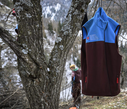 Affaiblissement du service civil : une attaque frontale de la droite Un gilet du Service civil suisse (bicolore bleu et bordeaux) accroché à un arbre en forêt, avec en arrière-plan une personne travaillant dans un paysage hivernal de montagne.