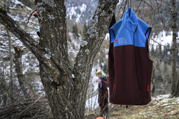 Un gilet du Service civil suisse (bicolore bleu et bordeaux) accroché à un arbre en forêt, avec en arrière-plan une personne travaillant dans un paysage hivernal de montagne.