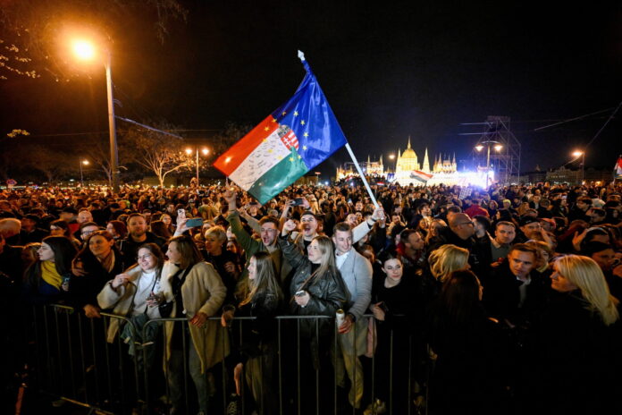 Rassemblement nocturne de milliers de personnes devant le Parlement hongrois à Budapest. Au centre, une personne brandit un drapeau hongrois combiné avec le drapeau de l'Union européenne. La foule, éclairée par des lampadaires, se tient derrière des barrières de sécurité. L'édifice du Parlement illuminé en arrière-plan. Manifestation politique contre Viktor Orbán.