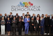 Andreas Missbach : « Avancer ensemble, maintenant » Une photo de groupe de personnalités politiques et de dirigeants internationaux, debout sur une estrade, posent devant une bannière blanche avec le slogan "En defensa de la democracia" (En défense de la démocratie) accompagné d'un logo coloré composé de cercles multicolores. Les participants, vêtus de tenues formelles, applaudissent, exprimant leur engagement et leur soutien à la démocratie. L'image capture un moment de solidarité et de collaboration internationale.