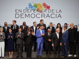 Andreas Missbach : « Avancer ensemble, maintenant » Une photo de groupe de personnalités politiques et de dirigeants internationaux, debout sur une estrade, posent devant une bannière blanche avec le slogan "En defensa de la democracia" (En défense de la démocratie) accompagné d'un logo coloré composé de cercles multicolores. Les participants, vêtus de tenues formelles, applaudissent, exprimant leur engagement et leur soutien à la démocratie. L'image capture un moment de solidarité et de collaboration internationale.
