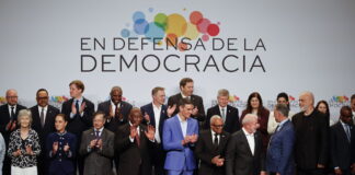 Andreas Missbach : « Avancer ensemble, maintenant » Une photo de groupe de personnalités politiques et de dirigeants internationaux, debout sur une estrade, posent devant une bannière blanche avec le slogan "En defensa de la democracia" (En défense de la démocratie) accompagné d'un logo coloré composé de cercles multicolores. Les participants, vêtus de tenues formelles, applaudissent, exprimant leur engagement et leur soutien à la démocratie. L'image capture un moment de solidarité et de collaboration internationale.