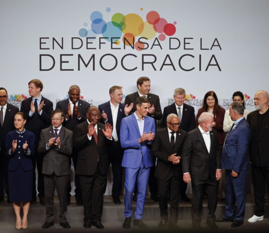 Andreas Missbach : « Avancer ensemble, maintenant » Une photo de groupe de personnalités politiques et de dirigeants internationaux, debout sur une estrade, posent devant une bannière blanche avec le slogan "En defensa de la democracia" (En défense de la démocratie) accompagné d'un logo coloré composé de cercles multicolores. Les participants, vêtus de tenues formelles, applaudissent, exprimant leur engagement et leur soutien à la démocratie. L'image capture un moment de solidarité et de collaboration internationale.