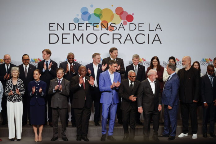 Une photo de groupe de personnalités politiques et de dirigeants internationaux, debout sur une estrade, posent devant une bannière blanche avec le slogan "En defensa de la democracia" (En défense de la démocratie) accompagné d'un logo coloré composé de cercles multicolores. Les participants, vêtus de tenues formelles, applaudissent, exprimant leur engagement et leur soutien à la démocratie. L'image capture un moment de solidarité et de collaboration internationale.