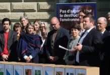 Initiative sur la place financière : 143 000 signatures déposées Une quinzaine de personnes, habillées en tenue formelle, sont rassemblées à l'extérieur d'un bâtiment institutionnel, vraisemblablement à Berne. Au premier plan, des boîtes portant des armoiries de cantons suisses sont alignées, suggérant un dépôt de signatures pour une initiative ou un référendum. En arrière-plan, une bannière bleue affiche le slogan « Pas d'argent pour la destruction de l'environnement ». Un homme tenant une feuille de papier semble prendre la parole devant le groupe.