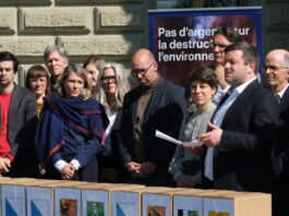 Initiative sur la place financière : 143 000 signatures déposées Une quinzaine de personnes, habillées en tenue formelle, sont rassemblées à l'extérieur d'un bâtiment institutionnel, vraisemblablement à Berne. Au premier plan, des boîtes portant des armoiries de cantons suisses sont alignées, suggérant un dépôt de signatures pour une initiative ou un référendum. En arrière-plan, une bannière bleue affiche le slogan « Pas d'argent pour la destruction de l'environnement ». Un homme tenant une feuille de papier semble prendre la parole devant le groupe.