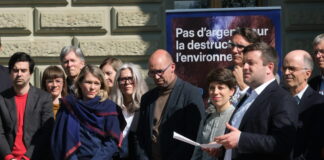 Initiative sur la place financière : 143 000 signatures déposées Une quinzaine de personnes, habillées en tenue formelle, sont rassemblées à l'extérieur d'un bâtiment institutionnel, vraisemblablement à Berne. Au premier plan, des boîtes portant des armoiries de cantons suisses sont alignées, suggérant un dépôt de signatures pour une initiative ou un référendum. En arrière-plan, une bannière bleue affiche le slogan « Pas d'argent pour la destruction de l'environnement ». Un homme tenant une feuille de papier semble prendre la parole devant le groupe.
