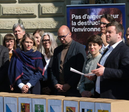Initiative sur la place financière : 143 000 signatures déposées Une quinzaine de personnes, habillées en tenue formelle, sont rassemblées à l'extérieur d'un bâtiment institutionnel, vraisemblablement à Berne. Au premier plan, des boîtes portant des armoiries de cantons suisses sont alignées, suggérant un dépôt de signatures pour une initiative ou un référendum. En arrière-plan, une bannière bleue affiche le slogan « Pas d'argent pour la destruction de l'environnement ». Un homme tenant une feuille de papier semble prendre la parole devant le groupe.