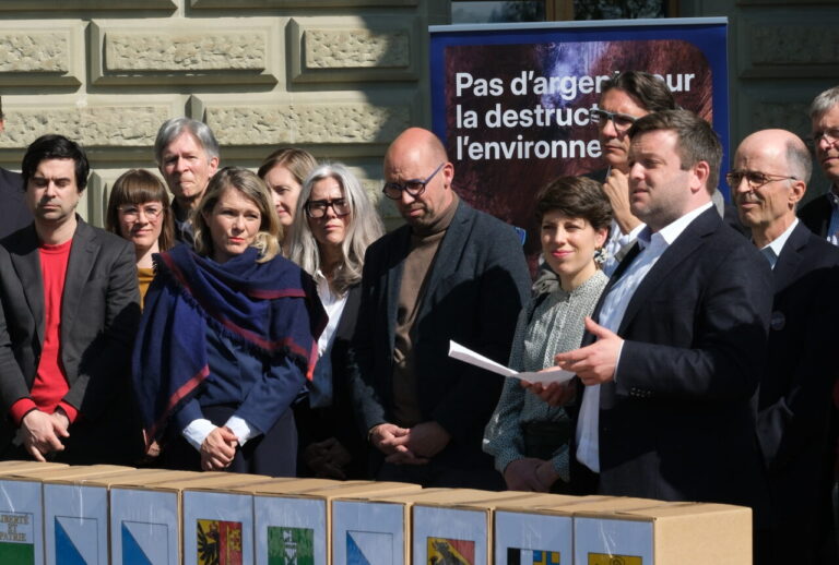 Une quinzaine de personnes, habillées en tenue formelle, sont rassemblées à l'extérieur d'un bâtiment institutionnel, vraisemblablement à Berne. Au premier plan, des boîtes portant des armoiries de cantons suisses sont alignées, suggérant un dépôt de signatures pour une initiative ou un référendum. En arrière-plan, une bannière bleue affiche le slogan « Pas d'argent pour la destruction de l'environnement ». Un homme tenant une feuille de papier semble prendre la parole devant le groupe.