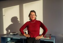 Blertë Berisha : « Depuis des années, nous constatons une fréquentation et une charge de travail constamment élevées dans les maisons d’accueil » foncé, portant un collier et un bracelet argentés. Elle pose les mains sur les hanches avec assurance devant un bureau bleu sur lequel reposent divers objets. En arrière-plan, un mur blanc est éclairé par la lumière naturelle, projetant son ombre de profil.