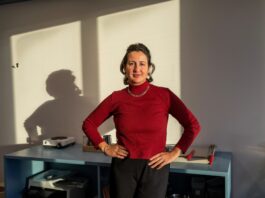 Blertë Berisha : « Depuis des années, nous constatons une fréquentation et une charge de travail constamment élevées dans les maisons d’accueil » foncé, portant un collier et un bracelet argentés. Elle pose les mains sur les hanches avec assurance devant un bureau bleu sur lequel reposent divers objets. En arrière-plan, un mur blanc est éclairé par la lumière naturelle, projetant son ombre de profil.