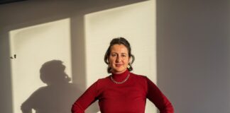 Blertë Berisha : « Depuis des années, nous constatons une fréquentation et une charge de travail constamment élevées dans les maisons d’accueil » foncé, portant un collier et un bracelet argentés. Elle pose les mains sur les hanches avec assurance devant un bureau bleu sur lequel reposent divers objets. En arrière-plan, un mur blanc est éclairé par la lumière naturelle, projetant son ombre de profil.