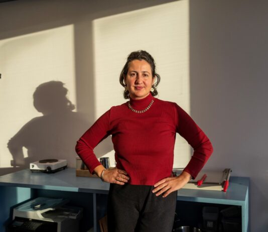 Blertë Berisha : « Depuis des années, nous constatons une fréquentation et une charge de travail constamment élevées dans les maisons d’accueil » foncé, portant un collier et un bracelet argentés. Elle pose les mains sur les hanches avec assurance devant un bureau bleu sur lequel reposent divers objets. En arrière-plan, un mur blanc est éclairé par la lumière naturelle, projetant son ombre de profil.