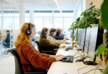 La Suisse, un pays peu favorable aux mères qui travaillent Un open space de bureau lumineux et moderne dans lequel plusieurs femmes sont assises en rangée le long d'un bureau blanc, portant des casques audio et travaillant sur des ordinateurs à double écran. L'espace est agrémenté de plantes vertes et bénéficie d'une lumière naturelle provenant de grandes fenêtres. Des cloisons en verre sont visibles en arrière-plan.