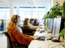 La Suisse, un pays peu favorable aux mères qui travaillent Un open space de bureau lumineux et moderne dans lequel plusieurs femmes sont assises en rangée le long d'un bureau blanc, portant des casques audio et travaillant sur des ordinateurs à double écran. L'espace est agrémenté de plantes vertes et bénéficie d'une lumière naturelle provenant de grandes fenêtres. Des cloisons en verre sont visibles en arrière-plan.