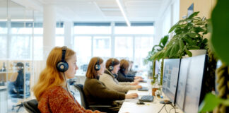 La Suisse, un pays peu favorable aux mères qui travaillent Un open space de bureau lumineux et moderne dans lequel plusieurs femmes sont assises en rangée le long d'un bureau blanc, portant des casques audio et travaillant sur des ordinateurs à double écran. L'espace est agrémenté de plantes vertes et bénéficie d'une lumière naturelle provenant de grandes fenêtres. Des cloisons en verre sont visibles en arrière-plan.