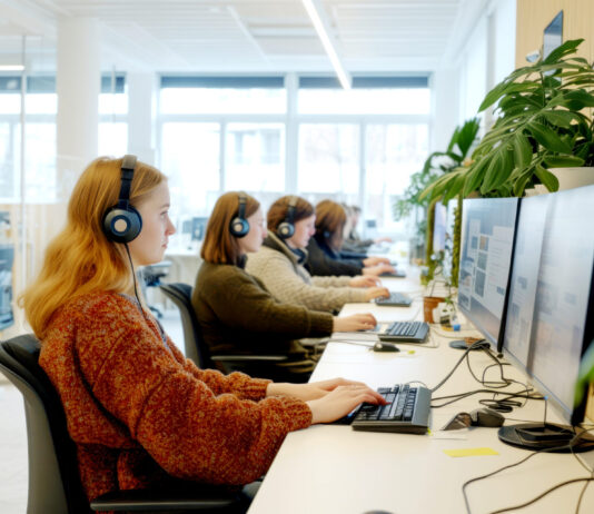 La Suisse, un pays peu favorable aux mères qui travaillent Un open space de bureau lumineux et moderne dans lequel plusieurs femmes sont assises en rangée le long d'un bureau blanc, portant des casques audio et travaillant sur des ordinateurs à double écran. L'espace est agrémenté de plantes vertes et bénéficie d'une lumière naturelle provenant de grandes fenêtres. Des cloisons en verre sont visibles en arrière-plan.