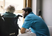 Nouvelle loi sur le service civil : quelles conséquences pour les EMS ? Un jeune homme en uniforme du service civil bleu se penche vers une personne âgée aux cheveux blancs assise dans un fauteuil roulant de marque Sopur. Ils sont face à une fenêtre dans un intérieur lumineux.