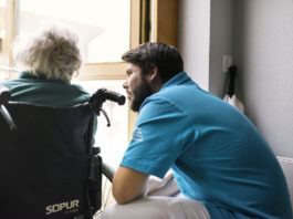 Nouvelle loi sur le service civil : quelles conséquences pour les EMS ? Un jeune homme en uniforme du service civil bleu se penche vers une personne âgée aux cheveux blancs assise dans un fauteuil roulant de marque Sopur. Ils sont face à une fenêtre dans un intérieur lumineux.