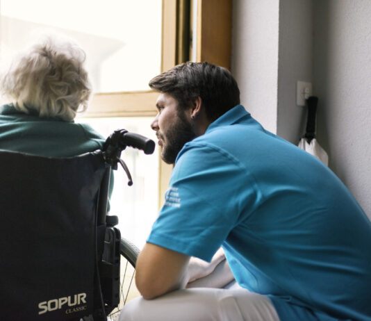 Nouvelle loi sur le service civil : quelles conséquences pour les EMS ? Un jeune homme en uniforme du service civil bleu se penche vers une personne âgée aux cheveux blancs assise dans un fauteuil roulant de marque Sopur. Ils sont face à une fenêtre dans un intérieur lumineux.