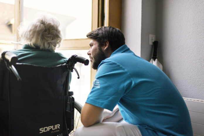 Un jeune homme en uniforme du service civil bleu se penche vers une personne âgée aux cheveux blancs assise dans un fauteuil roulant de marque Sopur. Ils sont face à une fenêtre dans un intérieur lumineux.