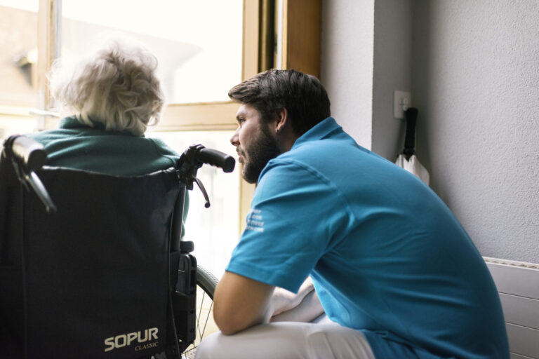 Un jeune homme en uniforme du service civil bleu se penche vers une personne âgée aux cheveux blancs assise dans un fauteuil roulant de marque Sopur. Ils sont face à une fenêtre dans un intérieur lumineux.