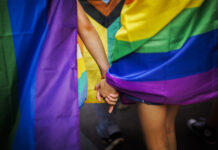 Queerophobie en Suisse : 80 % des personnes LGBTIQ sont victimes de violence Deux personnes se tenant la main, entourées de drapeaux aux couleurs arc-en-ciel lors d'une Pride (marche des fiertés LGBTIQ). On distingue notamment le drapeau Progress Pride, aux couleurs rouge, orange, jaune, vert, bleu, violet, blanc, rose et noir. L'image est cadrée sur les mains enlacées et les drapeaux colorés, créant une atmosphère de solidarité et de célébration.