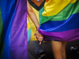 Queerophobie en Suisse : 80 % des personnes LGBTIQ sont victimes de violence Deux personnes se tenant la main, entourées de drapeaux aux couleurs arc-en-ciel lors d'une Pride (marche des fiertés LGBTIQ). On distingue notamment le drapeau Progress Pride, aux couleurs rouge, orange, jaune, vert, bleu, violet, blanc, rose et noir. L'image est cadrée sur les mains enlacées et les drapeaux colorés, créant une atmosphère de solidarité et de célébration.