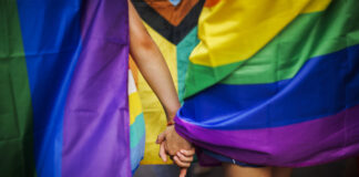Queerophobie en Suisse : 80 % des personnes LGBTIQ sont victimes de violence Deux personnes se tenant la main, entourées de drapeaux aux couleurs arc-en-ciel lors d'une Pride (marche des fiertés LGBTIQ). On distingue notamment le drapeau Progress Pride, aux couleurs rouge, orange, jaune, vert, bleu, violet, blanc, rose et noir. L'image est cadrée sur les mains enlacées et les drapeaux colorés, créant une atmosphère de solidarité et de célébration.