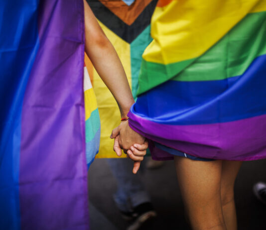 Queerophobie en Suisse : 80 % des personnes LGBTIQ sont victimes de violence Deux personnes se tenant la main, entourées de drapeaux aux couleurs arc-en-ciel lors d'une Pride (marche des fiertés LGBTIQ). On distingue notamment le drapeau Progress Pride, aux couleurs rouge, orange, jaune, vert, bleu, violet, blanc, rose et noir. L'image est cadrée sur les mains enlacées et les drapeaux colorés, créant une atmosphère de solidarité et de célébration.