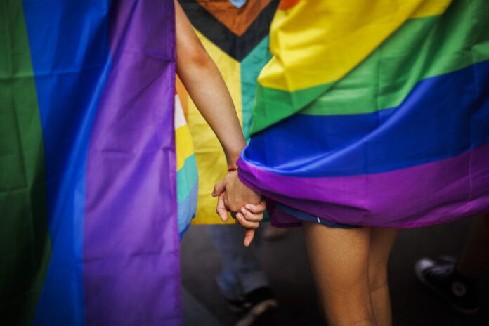 KUNDGEBUNG, EHE FUER ALLE, GLEICHGESCHLECHTLICHE EHE, LGBTIQ, LGBTIQ COMMUNITY, TRANSSEXUALITAET, ZURICH, Deux personnes se tenant la main, entourées de drapeaux aux couleurs arc-en-ciel lors d'une Pride (marche des fiertés LGBTIQ). On distingue notamment le drapeau Progress Pride, aux couleurs rouge, orange, jaune, vert, bleu, violet, blanc, rose et noir. L'image est cadrée sur les mains enlacées et les drapeaux colorés, créant une atmosphère de solidarité et de célébration.