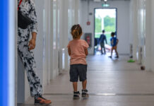 Avec son initiative, l’UDC met en péril les droits de l’enfant Photographie couleur : jeune enfant vue de dos, portant un t-shirt rose pêche et un short noir, marchant aux côtés d'un adulte en pantalon blanc imprimé et chaussures marron. Ils avancent dans un long couloir blanc aux murs lisses, éclairé naturellement. En arrière-plan flou, d'autres enfants jouent près d'une porte-fenêtre donnant sur l'extérieur vert.