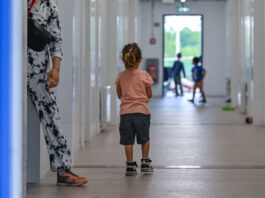 Avec son initiative, l’UDC met en péril les droits de l’enfant Photographie couleur : jeune enfant vue de dos, portant un t-shirt rose pêche et un short noir, marchant aux côtés d'un adulte en pantalon blanc imprimé et chaussures marron. Ils avancent dans un long couloir blanc aux murs lisses, éclairé naturellement. En arrière-plan flou, d'autres enfants jouent près d'une porte-fenêtre donnant sur l'extérieur vert.