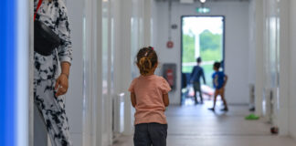Avec son initiative, l’UDC met en péril les droits de l’enfant Photographie couleur : jeune enfant vue de dos, portant un t-shirt rose pêche et un short noir, marchant aux côtés d'un adulte en pantalon blanc imprimé et chaussures marron. Ils avancent dans un long couloir blanc aux murs lisses, éclairé naturellement. En arrière-plan flou, d'autres enfants jouent près d'une porte-fenêtre donnant sur l'extérieur vert.