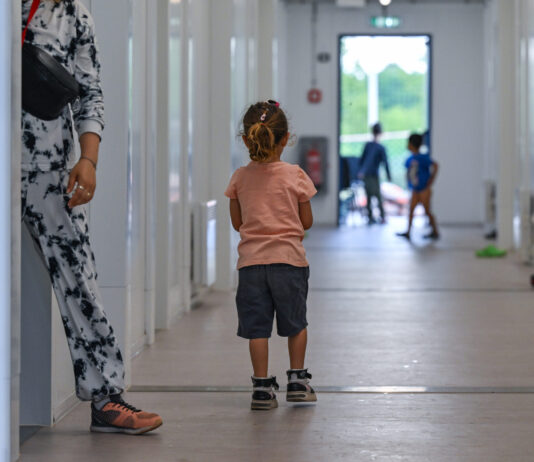Avec son initiative, l’UDC met en péril les droits de l’enfant Photographie couleur : jeune enfant vue de dos, portant un t-shirt rose pêche et un short noir, marchant aux côtés d'un adulte en pantalon blanc imprimé et chaussures marron. Ils avancent dans un long couloir blanc aux murs lisses, éclairé naturellement. En arrière-plan flou, d'autres enfants jouent près d'une porte-fenêtre donnant sur l'extérieur vert.