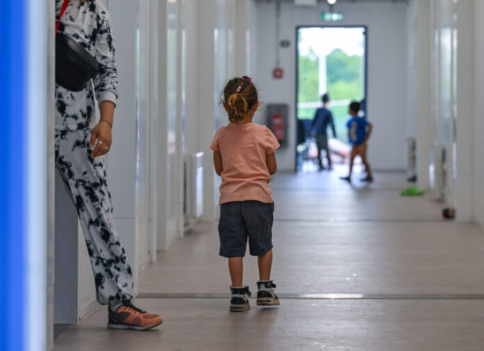 Photographie couleur : jeune enfant vue de dos, portant un t-shirt rose pêche et un short noir, marchant aux côtés d'un adulte en pantalon blanc imprimé et chaussures marron. Ils avancent dans un long couloir blanc aux murs lisses, éclairé naturellement. En arrière-plan flou, d'autres enfants jouent près d'une porte-fenêtre donnant sur l'extérieur vert.