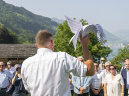 14 juin : le programme extrême de l’UDC pourrait devenir réalité Rassemblement de l'UDC sur la prairie du Grutli : Un homme en chemise blanche tient un modèle de planeur blanc levé vers le ciel, tandis qu'il se tient devant un groupe de personnes près d'un lac de montagne. En arrière-plan, on voit des montagnes boisées, des chalets traditionnels et un lac scintillant.