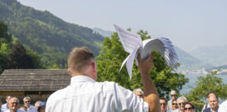 14 juin : le programme extrême de l’UDC pourrait devenir réalité Rassemblement de l'UDC sur la prairie du Grutli : Un homme en chemise blanche tient un modèle de planeur blanc levé vers le ciel, tandis qu'il se tient devant un groupe de personnes près d'un lac de montagne. En arrière-plan, on voit des montagnes boisées, des chalets traditionnels et un lac scintillant.